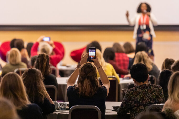 A woman speaking on stage and people watching and recording her on their phone.