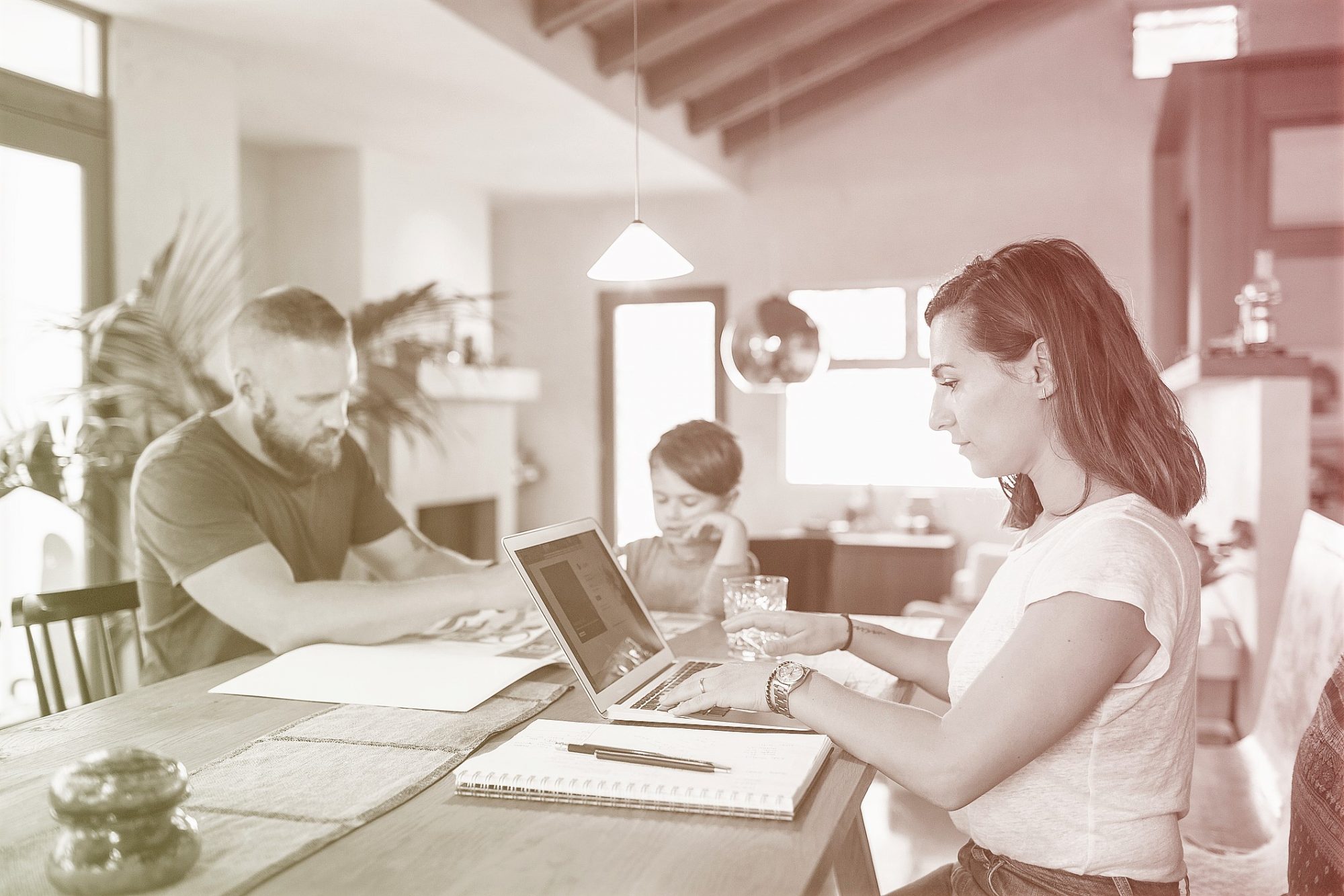 Woman working on laptop at dining table with family in background