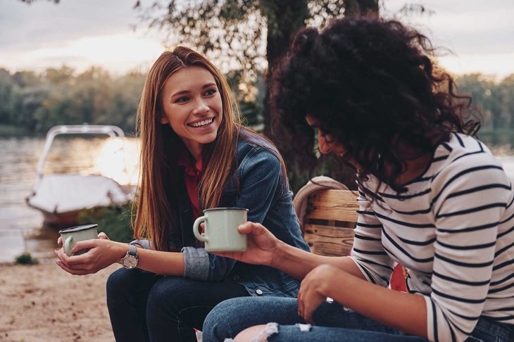 Good talk with friend. Two young beautiful women in casual wear smiling and talking while enjoying camping near the lake